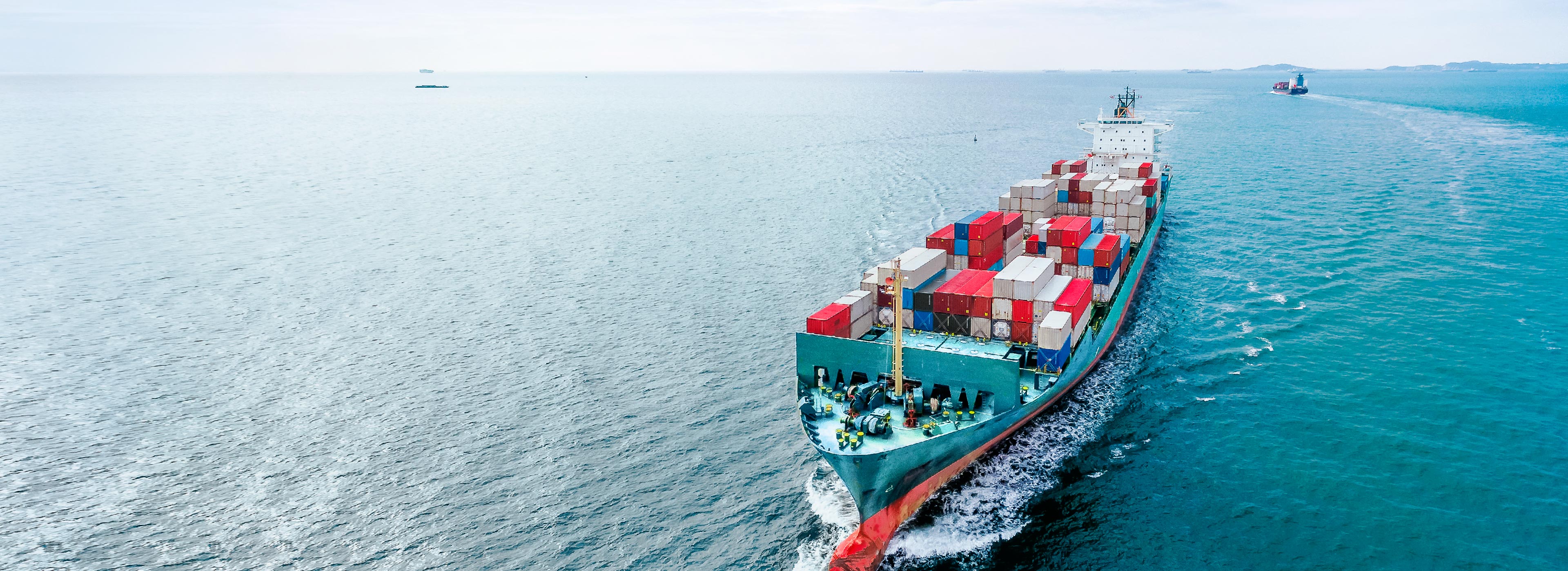 A cargo ship laden with colorful shipping containers sails through calm, blue waters. The vessel moves steadily, leaving a wake behind as it navigates the open sea. The horizon shows faint outlines of distant land and other ships in the background.