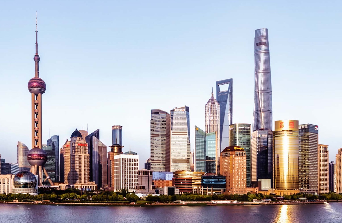 A panoramic view of the Shanghai skyline features iconic skyscrapers, including the Oriental Pearl Tower and the Shanghai Tower. The buildings reflect modern architecture against a clear sky, showcasing the vibrant urban landscape along the riverbank.