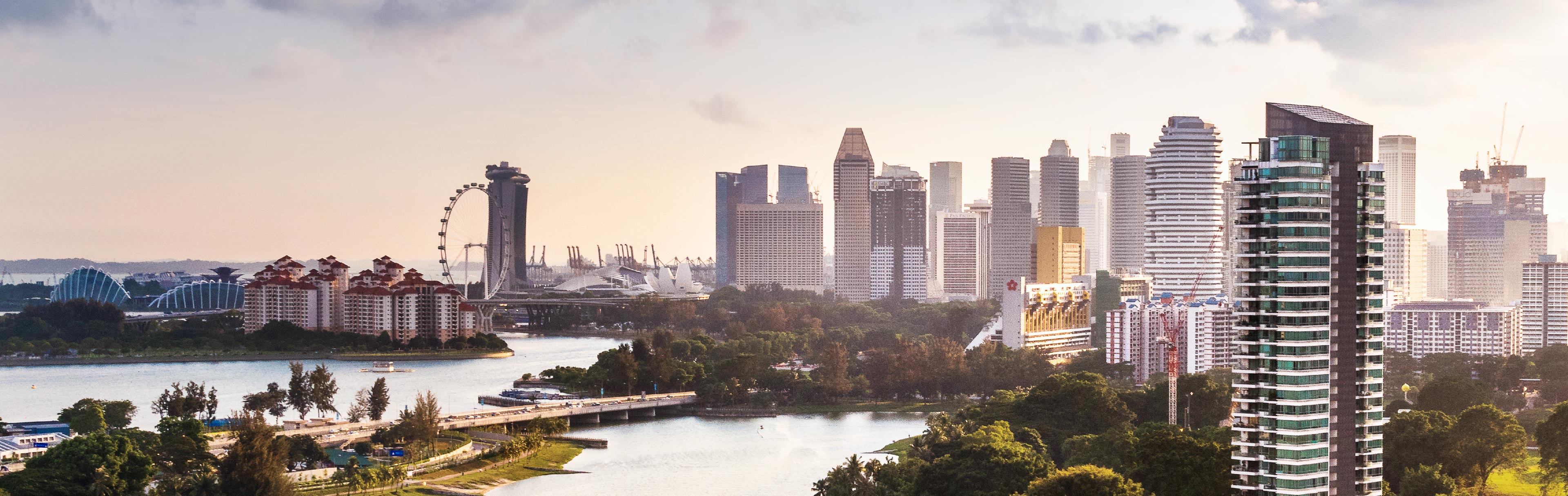 A panoramic view of a modern city skyline featuring tall skyscrapers, residential buildings, and a large Ferris wheel. In the foreground, a river flows through green parks, with a bridge crossing it. The scene is illuminated by soft sunlight, creating a tranquil atmosphere.