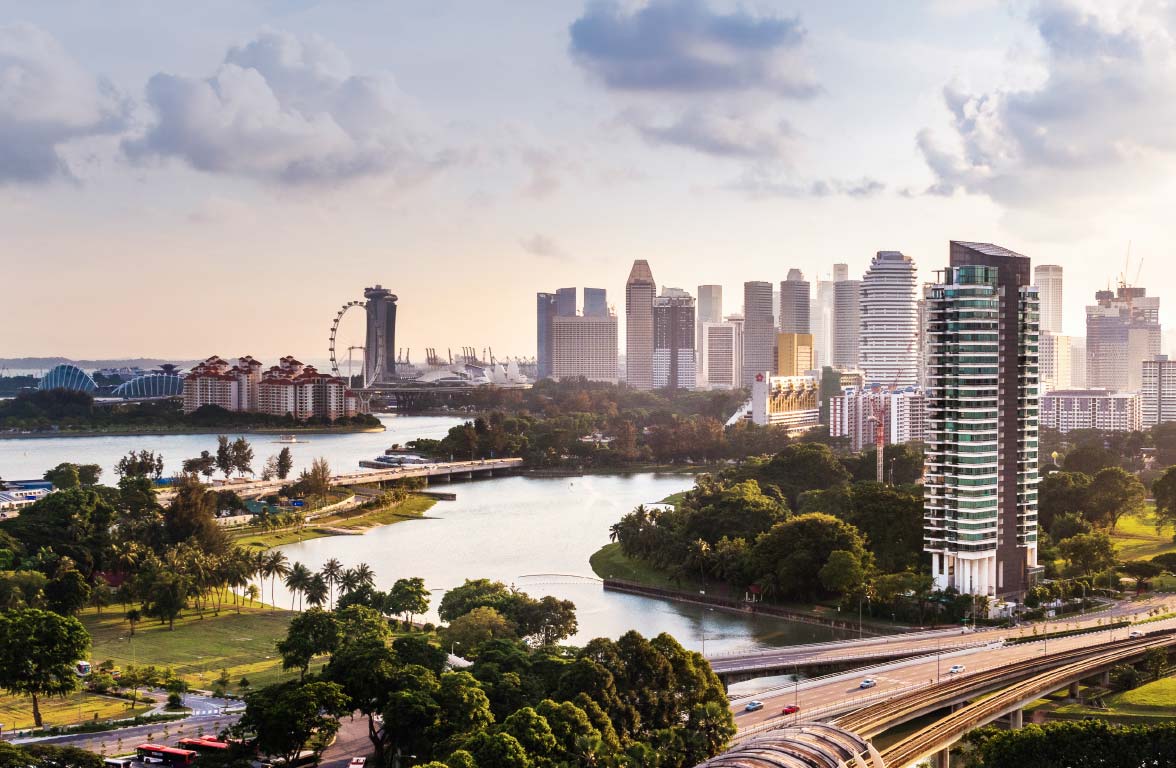 A panoramic view of a modern city skyline at sunset, featuring skyscrapers, a large wheel, and lush greenery along a serene waterfront with gently flowing water. Roads weave through the landscape, highlighting a blend of urban and natural elements.