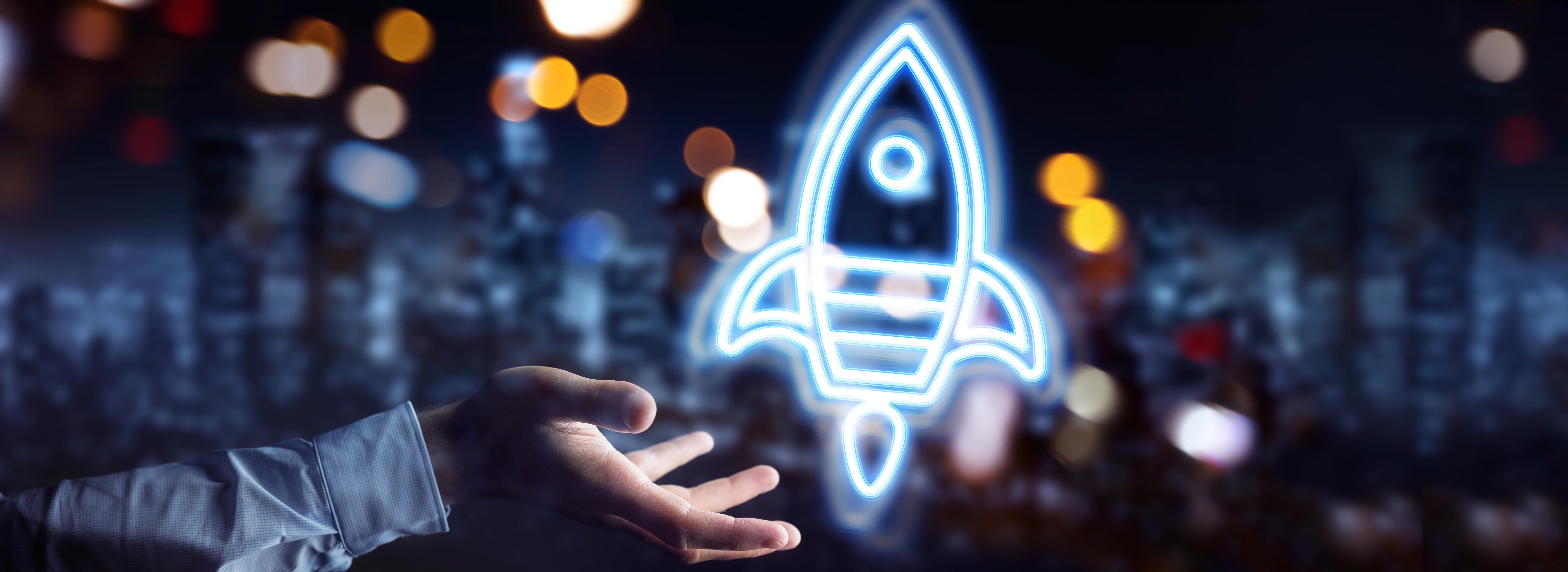 A hand reaches out towards a glowing blue rocket icon, symbolizing innovation and new ideas. The background is blurred, suggesting a dynamic environment. This image conveys themes of progress and exploration in technology or entrepreneurship.