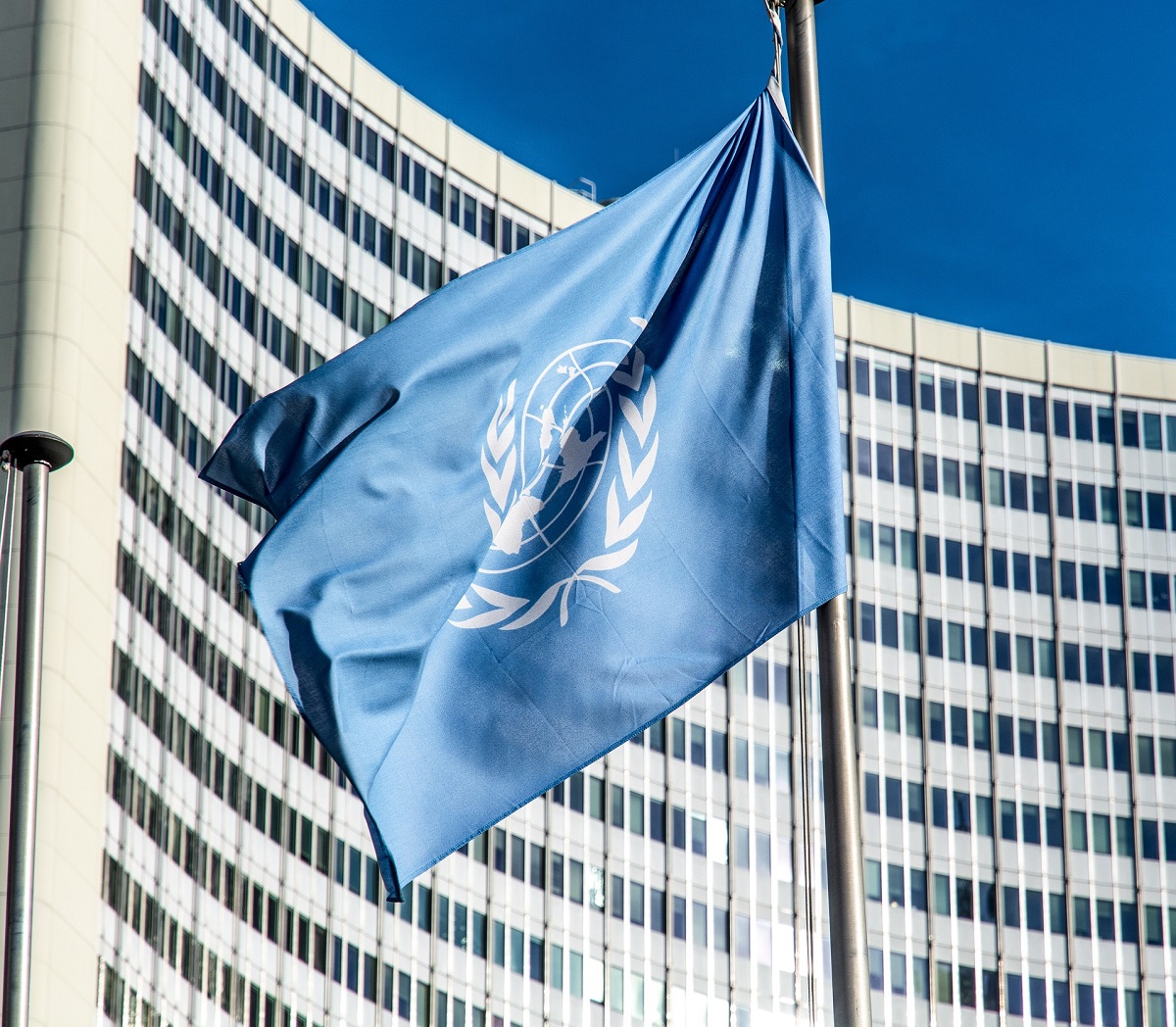 A blue flag with the emblem of the United Nations, featuring a world map and olive branches, is waving. In the background, a modern, curved building with many windows is visible under a clear blue sky.