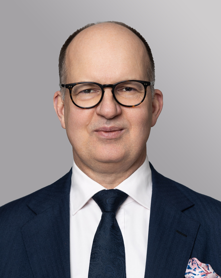 Ein Mann mit Brille trägt einen dunkelblauen Anzug und ein weißes Hemd. Er hat einen dunklen Krawatte mit Muster und ein pinkfarbenes Taschentuch in der Jackettasche. Der Hintergrund ist grau. Der Mann lächelt leicht und schaut direkt in die Kamera.